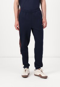 Marineblå sportsbukser med elastisk talje, sidestriber i maroon og creme, og glat stof. Sammen med beige sneakers.