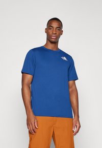 Μπλέ κοντομάνικο μπλουζάκι από λείο ύφασμα, με μικρό λευκό λογότυπο στο αριστερό στήθος. Συνδυάζεται με πορτοκαλί σορτς.