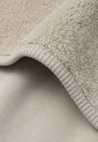 Beige fleece deken met een gestructureerd, pluizig oppervlak en een gladde achterkant. Voorzien van een geribbelde randafwerking, zacht en warm uiterlijk.