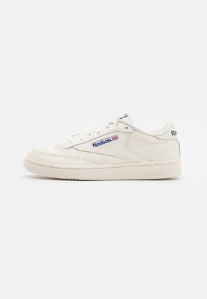 Biały niski sneaker Reebok z niebieskim logo i emblem emblemem brytyjskiej flagi, białe sznurowadła oraz teksturowana gumowa podeszwa na jednolitym tle.