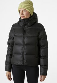 Schwarze Steppjacke mit hohem Kragen, ausgestattet mit einem Frontreißverschluss und einer einzelnen Tasche. Getragen mit einer gelben gerippten Strickmütze.