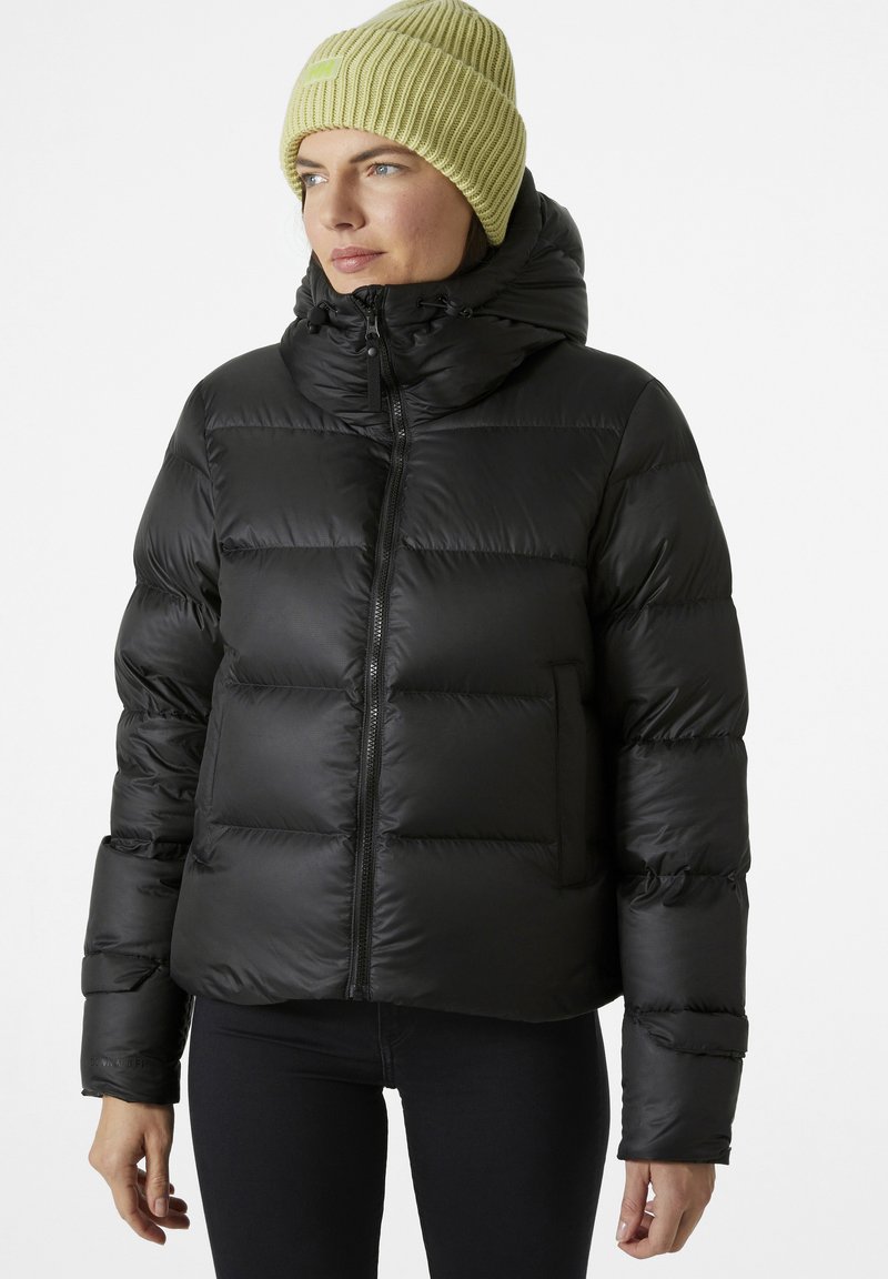Schwarze Steppjacke mit hohem Kragen, ausgestattet mit einem Frontreißverschluss und einer einzelnen Tasche. Getragen mit einer gelben gerippten Strickmütze.