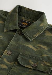 Chemise boutonnée vert camouflage en tissu durable, avec un col classique, deux poches poitrine et des boutons marron. Étiquette de marque visible à l'intérieur.