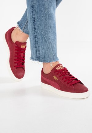 Person iført røde Puma-sneakers med tekstureret design og guldlogo, kombineret med blå jeans med frynset kant mod en hvid baggrund.