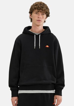 Joven con cabello rizado de pie mirando al frente, lleva una sudadera negra con capucha y un pequeño logotipo rojo y amarillo en el pecho.