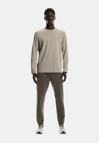 Top bej cu mâneci lungi și pantaloni maro deschis, confecționați dintr-un material neted. Modelul poartă adidași albi cu accente negre. Design minimalist.