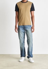 Men's t-shirt with brown front and black sleeves, featuring a small green logo; paired with light blue jeans and white sneakers.