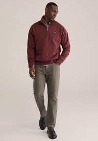 Tmavohnedá mikina na zips s rebrovanými manžetami, kombinovaná so sivozelenými nohavicami a čiernymi koženými topánkami. Na hrudi je malá značka.