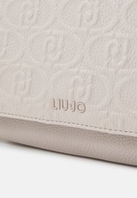 Beżowa torebka ze skóry o teksturowanej powierzchni z tłoczonym wzorem i wypukłym logo, z delikatnym przeszyciem i gładkim wykończeniem.