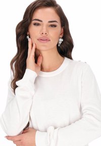 Witte top met lange mouwen van zachte stof, voorzien van geribbelde manchetten en decoratieve knopen op de schouder. Model draagt opvallende oorbellen.