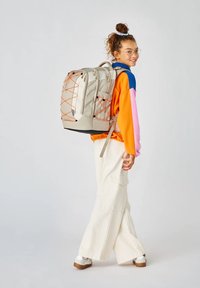 Helles grauer Rucksack mit orangefarbenen Akzenten, mehreren Fächern und einer strukturierten Oberfläche. Das Modell trägt einen mehrfarbigen Pullover und cremfarbene Hosen.