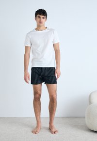 Fehér pamut póló fekete csíkos rövidnadrággal párosítva. A modell mezítláb áll egy világos szőnyegen, egy sima háttér előtt.