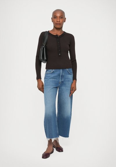 Top marrón de manga larga acanalada con una tapeta de botones, combinado con jeans de pierna ancha en azul claro con dobladillos deshilachados y bailarinas burdeos. Bolso negro.