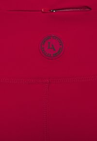 Vêtement de sport rouge avec une texture lisse et extensible, présentant un logo de marque circulaire en noir et un petit détail de poche zippée.