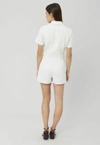 Short en tissu blanc texturé avec des manches courtes, un col, une taille cintrée et des poches arrière, associé à des chaussures à talons marron.