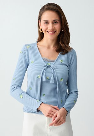Vrouw glimlacht, draagt een lichtblauw geribd vest met kleine gele bloemenpatronen en een witte broek, staand tegen een effen achtergrond.