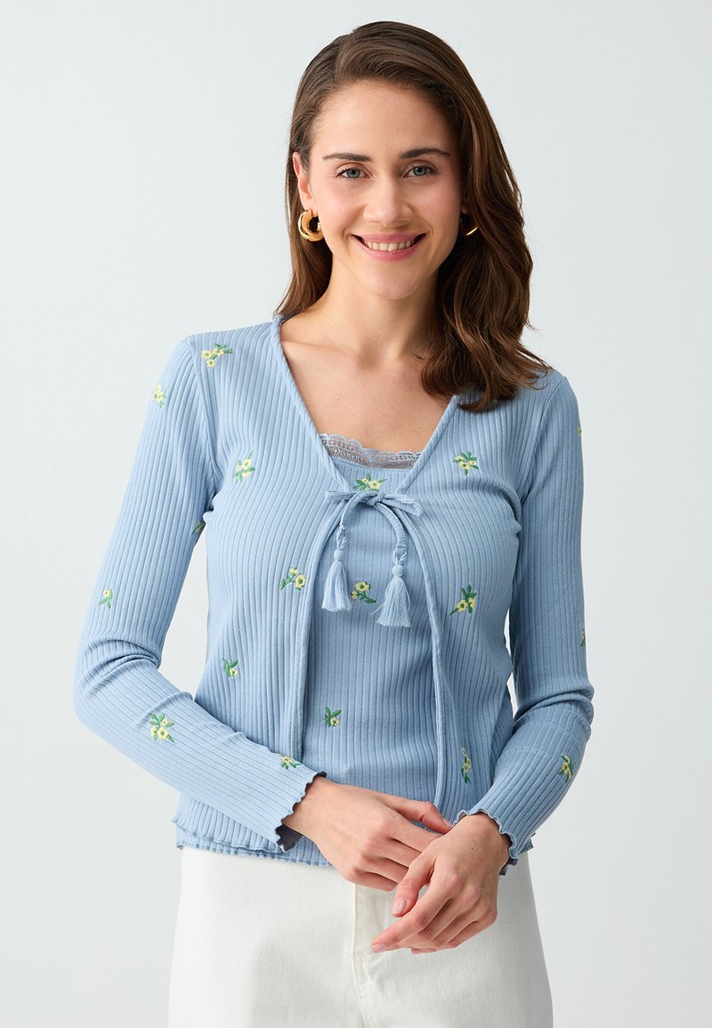 Femme souriante, portant un cardigan en maille côtelée bleu clair avec de petits motifs floraux jaunes et un pantalon blanc, debout devant un fond uni.