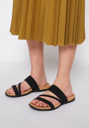 Chanclas negras con tres correas de tela, plantilla de corcho y suela de goma. Colocadas al lado de una falda de color mostaza con pliegues.