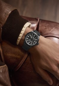 Sort armbåndsur med en rund urskive, hvide tal og silikonesnor. Båret på en hånd mod en brun læderbaggrund.