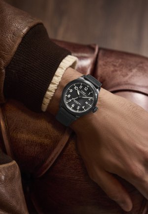 Schwarze Armbanduhr mit rundem Ziffernblatt, weißen Ziffern und Silikonarmband. Am Handgelenk vor einem braunen Lederhintergrund getragen.