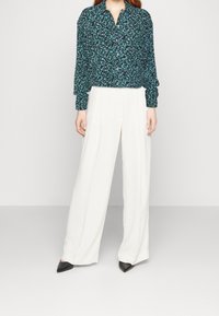 Long-sleeve, collared blouse in dark green with a floral pattern paired with high-waisted, wide-leg white trousers and black pointed-toe heels.