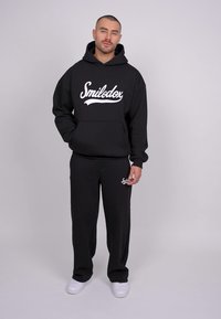 Sort hoodie med "Smilodxx" logo i hvid, parret med matchende sorte sweatpants. Glat stof, afslappet pasform og hvide sneakers.