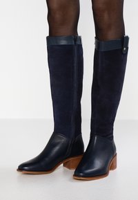 Granatowe kozaki za kolano z zamszowej cholewki, skórzanym czubkiem i drewnianą podeszwą. Posiadają zamek z boku oraz kontrastującą skórzaną taśmę na górze.