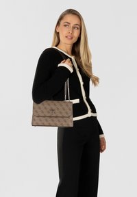 Borsa a tracolla marrone con motivo e catena. Il logo è ben visibile sul davanti. La borsa ha una forma rettangolare elegante e una finitura testurizzata.