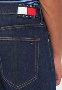 Tumed denim püksid on varustatud tagakotikaga, millel on punane, valge ja sinine logoäkk ning kontrastne kollane õmblus tasku servadel.