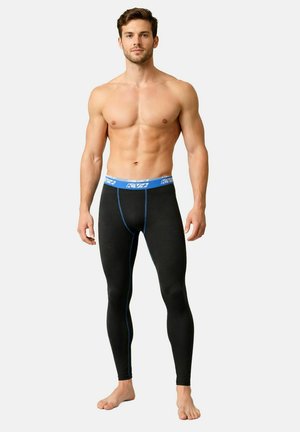 Uomo muscoloso in piedi a piedi nudi, indossa leggings aderenti neri con vita e cuciture blu, su uno sfondo chiaro uniforme.