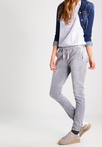 Pantalon de jogging en tissu gris doux avec une ceinture élastique, doté de poches latérales. Porté avec une chemise claire à boutons et une veste en denim.
