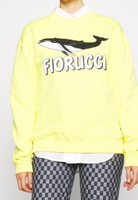 Sudadera amarilla brillante con un gráfico de ballena en blanco y negro, con el texto "Fiorucci" en una fuente bold y contrastante. Puños y dobladillo acanalados.