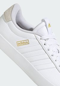 White leather sneaker with textured finish, featuring a gold "adidas" logo, white laces, and a light gray suede accent on the heel.
