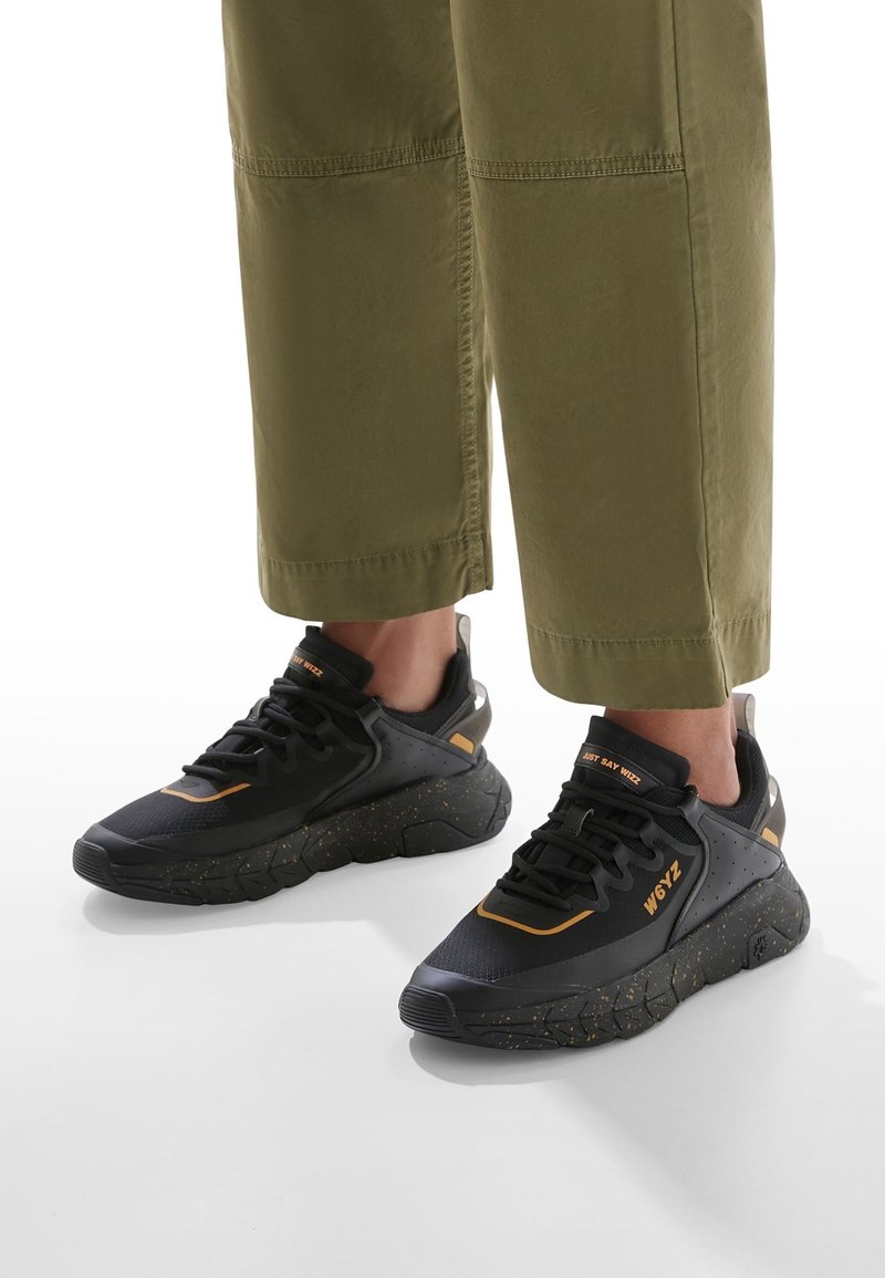 Baskets noires et orange avec des semelles épaisses tachetées portées avec un pantalon court vert olive sur fond blanc.