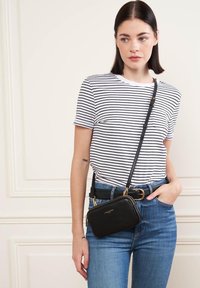 Borsa a tracolla in pelle nera con finitura texture, dettagli in hardware dorato e tracolla staccabile. Indossata sopra una maglietta a righe e jeans.