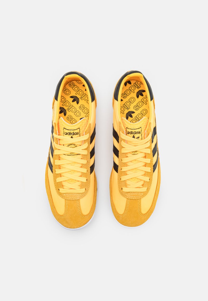 Gelbe Sneakers aus Stoff und Wildleder, mit schwarzen Streifen, runden Schnürsenkeln und einer gemusterten Innensohle mit "adidas" Branding.