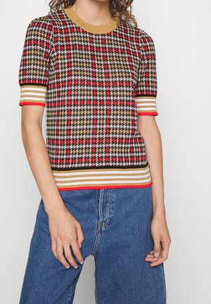 Personne portant un top en maille à manches courtes avec un motif pied-de-poule rouge, noir, blanc et or et une bordure rayée, associé à un jean bleu.