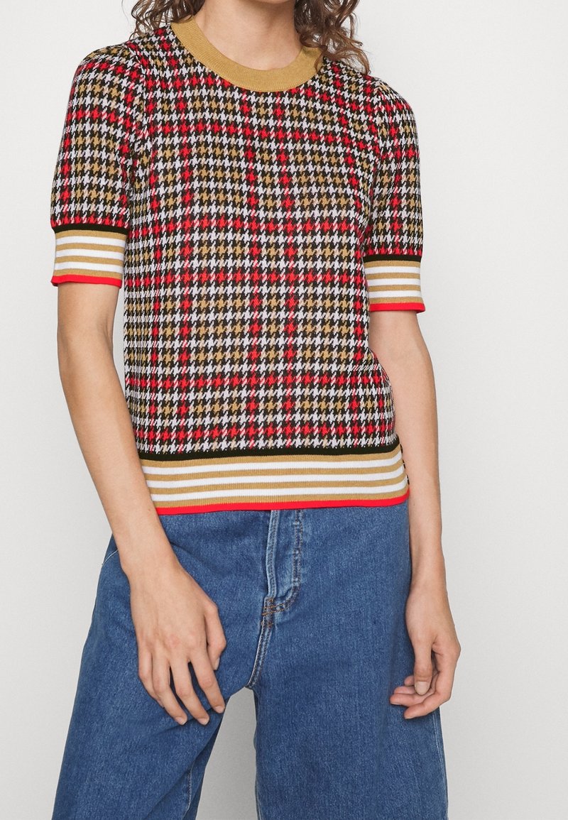 Personne portant un top en maille à manches courtes avec un motif pied-de-poule rouge, noir, blanc et or et une bordure rayée, associé à un jean bleu.