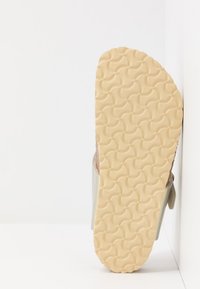 Sandal med en texturerad beige gummisula, med ett ljusgrått band och en brun fotbädd. Designen är platt med en enkel silhuett.