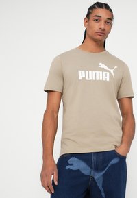 Bež pamukasta majica s kratkim rukavima i okruglim izrezom, s bijelim Puma logotipom na prednjoj strani. Model nosi plave traperice.