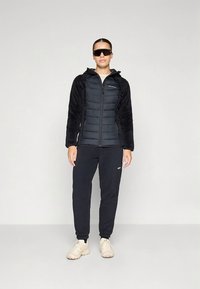 Sort dunjakke med bløde, sorte ærmer, lynlåslukning og logo på venstre bryst. Kombineret med mørke joggingbukser og lyse sko.