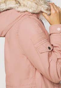 Růžová bunda parka s kapucí podšitou umělou kožešinou. Obsahuje manžety na knoflíky, boční kapsy a hladký materiál.