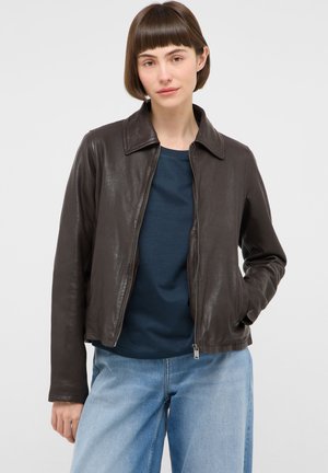 Frau mit kurzen braunen Haaren, die eine dunkelbraune Lederjacke über einem marineblauen Hemd und hellblauen Jeans trägt, eine Hand in der Tasche.