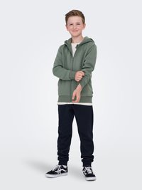 Felpa verde con zip, dal morbido tessuto, indossata sopra una camicia bianca. Abbinata a pantaloni della tuta blu scuro e scarpe nere con lacci bianchi.