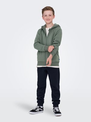 Hoodie zippé vert avec une texture douce, porté sur une chemise blanche. Associé à un pantalon de jogging bleu marine foncé et des baskets noires avec des lacets blancs.