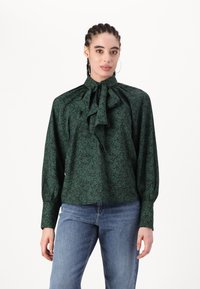 Blusa verde con motivo floreale, colletto alto e dettaglio a fiocco, maniche lunghe e tessuto arricciato, abbinata a jeans blu.