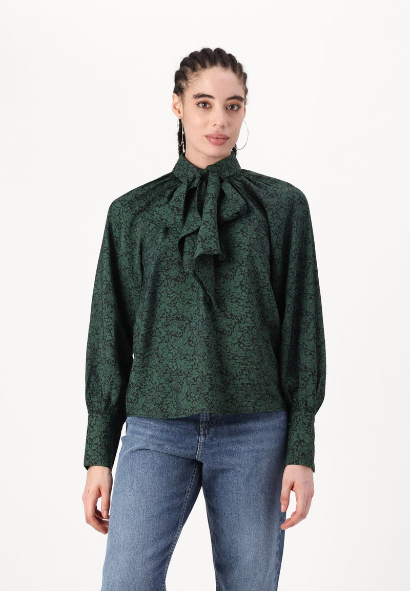 Blusa verde con motivo floreale, colletto alto e dettaglio a fiocco, maniche lunghe e tessuto arricciato, abbinata a jeans blu.