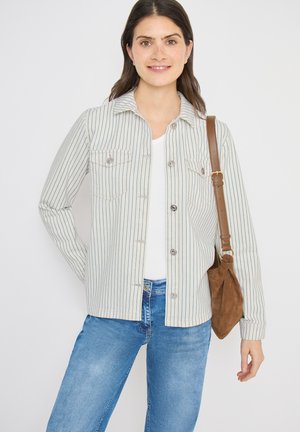 Femme souriante portant une veste blanche à fines rayures, un haut blanc, un jean bleu et un sac à bandoulière en daim marron, sur fond blanc.