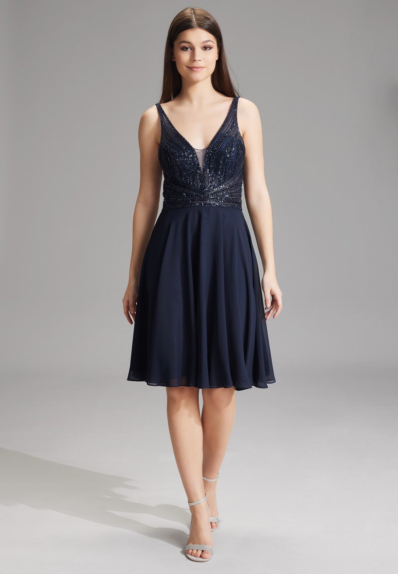 Abito A-line blu navy con corpetto aderente decorato con perline e scollatura a V. Gonna fluida realizzata in materiale leggero, abbinata a tacchi a stiletto argento.