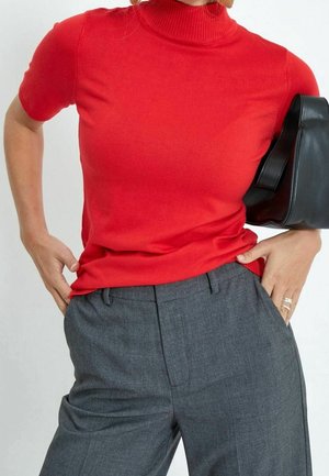 Persona vestida con una blusa roja de manga corta tipo cuello simulado y pantalones grises, sosteniendo un bolso negro bajo el brazo con las manos en las caderas.